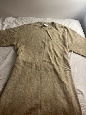 Beige stickad klänning med knyt - Mysig beige stickad klänning med rund halsringning och knytband i midjan för en snygg siluett. Klänningen har lång ärm och är perfekt för kyligare dagar. Enkel och stilren design som passar till många olika looks.