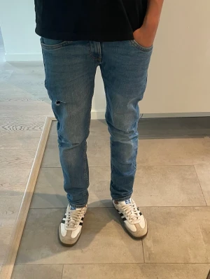 Blå slim fit jeans med slitning - Säljer ett par blå slim fit jeans i klassisk denim med en snygg slitning på vänster lår. Men det är 2 hål men folk kanske gillar det.