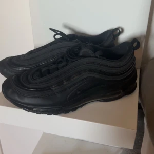 Helsvarta Nike Air Max 97 sneakers - Säljer ett par helsvarta Nike Air Max 97 sneakers med den ikoniska vågiga designen och synlig luftdämpning i sulan. Skorna har en låg silhuett, rund tå och snörning framtill. Materialet är en mix av textil och syntet för en sportig look.
