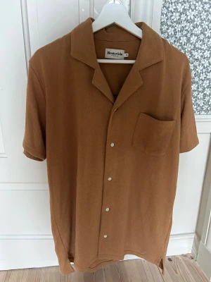 Brun skjorta från Resteröds - Säljer en brun skjorta från Resteröds i mjukt material. Endast testad. Skjortan har korta ärmar, klassisk krage, bröstficka och knappar framtill. Perfekt för dig som gillar en avslappnad och stilren look. 