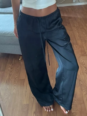 Svarta satinbyxor - Säljer ett par svarta, vida loungewear-byxor i glansigt satintyg. Byxorna har resår i midjan och dragsko för skön passform. Perfekta för chill dagar hemma eller när du vill vara extra bekväm.