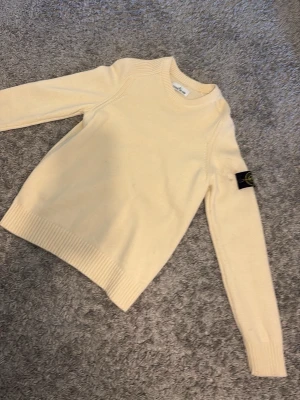 Beige stickad tröja från Stone Island - Snygg beige stickad tröja från Stone Island med klassisk rund halsringning och ribbade muddar. Tröjan har den ikoniska svarta logotypen med gult och grönt på vänster ärm. Perfekt för dig som gillar stilrena plagg med streetwear-vibe.