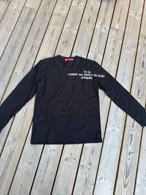 Svart långärmad Supreme x Comme des Garçons topp - Svart långärmad topp från Supreme i samarbete med Comme des Garçons. Tryck i vitt framtill med texten 'COMME des GARÇONS SHIRT SUPREME 15-16'. Klassisk rund hals och relaxed fit, perfekt för en clean streetwear-look.