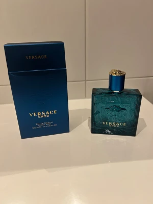 Versace Eros Eau de Toilette 100 ml - Versace Eros Eau de Toilette i en snygg turkosblå glasflaska med guldiga detaljer och Medusa-huvud på framsidan. Flaskan har en fyrkantig form och kommer med matchande blå kartong. Doften är maskulin och ikonisk.