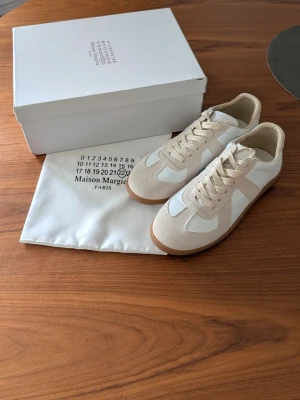 Maison Margiela vita och beige sneakers - Maison Margiela sneakers i vitt och beige med klassisk låg siluett. Ovandel i en mix av mocka och slätt läder, vita snören och gummisula. Snygg minimalistisk design som passar till det mesta. Kommer med originalkartong och dustbag.