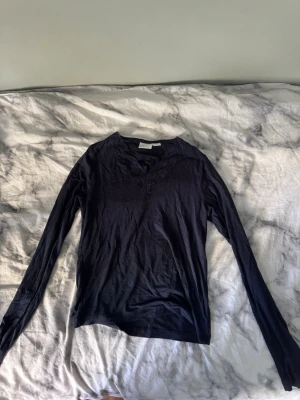 Svart långärmad topp med spets - Svart långärmad topp med v-ringning och dekorativ spetsdetalj runt halsen. Tunn och mjuk i materialet, perfekt för lager på lager eller som den är. Enkel och stilren design som passar till det mesta.