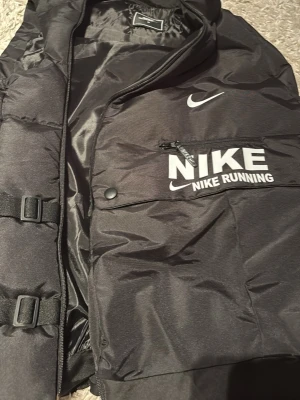 Svart dunväst från Nike Running - Snygg svart dunväst från Nike Running med vita loggor och dragkedja framtill. Västen har spännen på sidan och en ficka med dragkedja på bröstet. Perfekt för lager på lager och sportiga outfits.