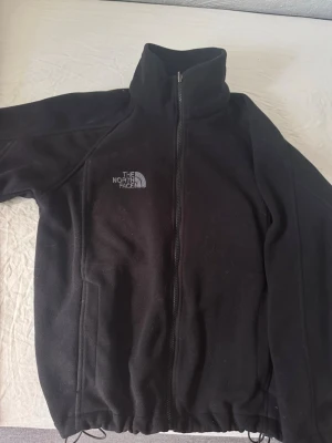 Svart fleecejacka från The North Face - Säljer en fodrad svart fleecejacka från The North Face med hel dragkedja och hög krage. Jackan har två sidofickor och broderad logga på bröstet. Perfekt för kyliga dagar och outdoor-aktiviteter. Tillverkad i mjukt fleecematerial.