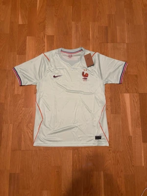 Vit fotbollströja Frankrike Nike - Snygg vit fotbollströja från Nike med Frankrikes landslagsemblem på bröstet. Tröjan har korta ärmar, rund hals och detaljer i rött och blått vid ärmslut och hals. Perfekt för dig som älskar fotboll och vill visa ditt stöd för Frankrike.