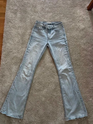 Ljusblåa utsvängda jeans - Säljer ett par ljusblå jeans med utsvängda ben och råa, fransiga benslut. Klassisk femficksmodell med mellan midja och dragkedja. Perfekta för dig som gillar retrostil och vill sticka ut lite extra.