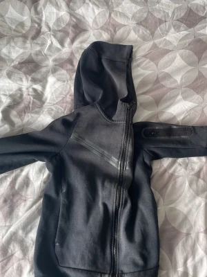 Svart sportig hoodie med dragkedja - Svart hoodie med hel dragkedja och huva. Jackan har två snedställda fickor framtill och diskreta reflexdetaljer på bröstet. Materialet ser ut att vara mjukt och stretchigt, perfekt för träning eller chill. Ärmen har en liten ficka med dragkedja för nycklar eller kort.
