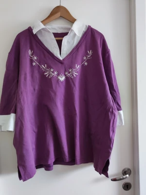 Lila oversized topp med broderi - Står ej storlek, men uppskattar att det är XL! Skriv om ni behöver fler mått:) Lila oversized topp med vit krage och manschetter som ger en lager-på-lager-look. Toppen har v-ringning och vitt broderat blommönster vid halsen. Perfekt för dig som gillar en avslappnad men ändå stilren vibe.