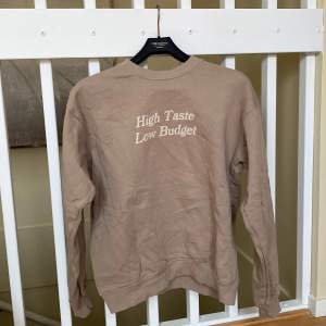 En ljusbrun sweatshirt från valient som är nästan helt oanvänd, inga skador eller liknande på tyget och är i nyskick! Med ett citat på som säger ”High taste low budget” Orginalpris: 350kr