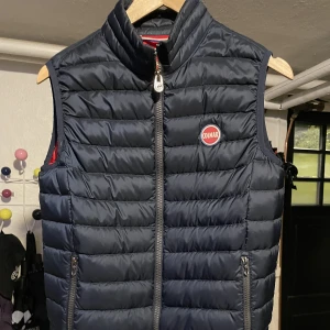 Colmar väst - Marinblå colmarväst med en liten skråma på vänster framsida men som annars är i bra skick. Se bild 2 för den lilla skadan.