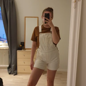 Vita korta snickarbyxor - Vita korta hängselbyxor som går bra att vika upp. Knappar på båda sidorna. Diskret distressed på framsidan. Använda en gång. Storleken är M men passar även S bra