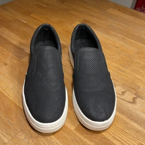 Svarta skor med hög sula - Svarta loafer-liknande skor med ormskinnsimitation. Tån är lite nött men annars finns inga tydliga skador. Lågt skaft. Ca 4cm hög sula. Storlek 41.