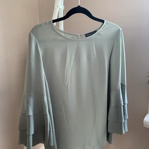 Ljusgrön blus med volangärm  - Superfin ljusgrön blus med volangärmar från STOCKHLM i storlek 36 som stängs baktill med en liten knapp. Vid granskning hittade jag en mycket liten tråddragning framtill på blusen, se bild. I övrigt inga anmärkningar.