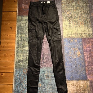 Tighta cargo skinnbyxor - Svarta byxor i skinnimmitation med fickor från Nelly.  Stl 36. Köparen står för frakt!