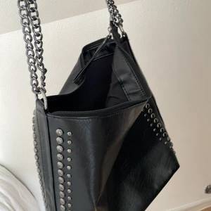 Väska - Svart väska i fejk skinn och mörka siver nitar. Handtaget är en kedja och en räm. Tre fack.