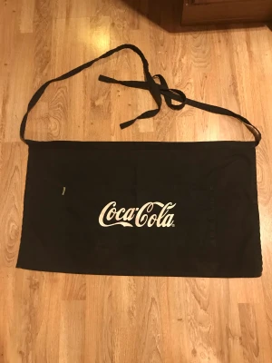Coca cola förkläde  - Svart midje förkläde i bomull. Coca cola text på framsidan. Två fickor. Snyggt i köket! 