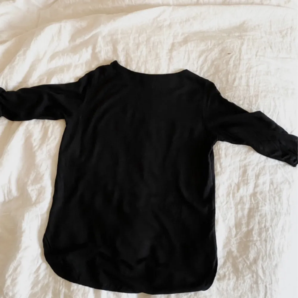 En svart blus med trekvartsärmar.  Det finns knappar på ärmarna som håller den uppvikta ärmen på plats.  (Se bild 2)  Mått:  - B: 40 cm - L: 65 cm. Blusar.