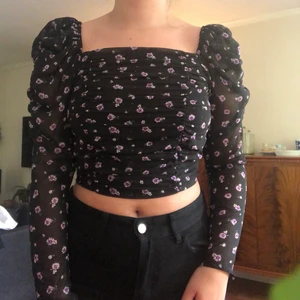 Blus med lila blommor!  - En kortare svart blus med lila blommor som man själv kan välja om man vill ha den off shoulder eller med ärmarna på. Aldrig använd! 