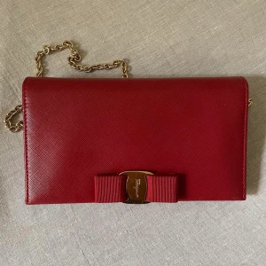 Salvatore Ferragamo Crossbody Wallet in Red Saffiano Leather - Red Saffiano Leather Embossed Logos Removable chain with clasps  Serial Number included  Interior 7 Card Insert, 2 Bill Inserts, Snap Flap Pocket,Removeable Gold Chain Strap Some marks in the hardware and lining Overall Very Good Made in Italy 19.5 x 11CM