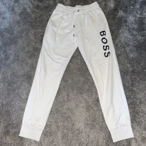 Vita mjukisbyxor från Boss - Säljer ett par vita mjukisbyxor från Boss. De har en elastisk midja med snörning och muddar vid bensluten. Perfekta för en avslappnad stil eller hemmamys. Boss-loggan är tryckt på ena benet för en stilren look.