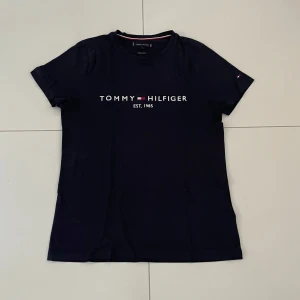 Mörkblå t-shirt från Tommy Hilfiger - Säljer en snygg mörkblå t-shirt från Tommy Hilfiger med deras ikoniska logga tryckt på framsidan. T-shirten är kortärmad och perfekt för en casual look. Passar både till vardags och fest. Skön och stilren design!