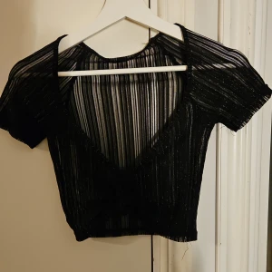 Svart glittrig topp nyskick  - Snygg svart glittrig topp med korta ärmar och djup v-ringning. Perfekt för festliga tillfällen! Materialet är lätt och transparent med ett subtilt randigt mönster. Toppen har en knytning framtill som ger en extra touch. Lappen är bortklippt tyvärr 