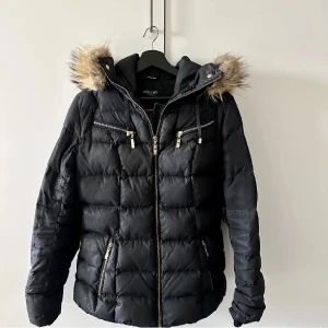 Hollies vinterjacka  - Svart jacka från Hollies med huva och äkta pälskant. Jackan har två fickor på sidorna och en innerficka. Jackan har även en dragkedja framtill. Saknar bälte.  Ordpris: 4500-5000kr. Passar M-XL. 