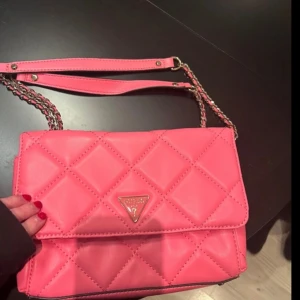 Rosa quiltad axelväska från Guess - Snygg rosa axelväska från Guess med quiltad design och kedjedetaljer på axelremmen. Väskan har en praktisk dragkedja inuti och stängs med tryckknappar. Perfekt för att ge en färgklick till din outfit! Säljs för den inte kommer till användning, nypris ca 2 000kr säljes för 1000kr 