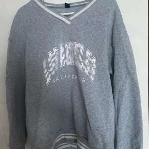 Grå sweatshirt med Los Angeles-tryck - Säljer en grå sweatshirt med texten 'Los Angeles California' i vitt på framsidan. Tröjan har en v-ringad design med randiga detaljer vid halsen och nederkanten. Perfekt för en avslappnad stil.