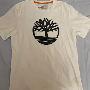 Vit t-shirt från Timberland - Snygg vit t-shirt från Timberland med deras ikoniska trädlogga i svart på bröstet. Tillverkad av 100% ekologisk bomull för en skön och hållbar känsla. Perfekt för en avslappnad stil.