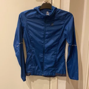 Blå vindjacka från Salomon - Säljer en blå superlätt o tunn vindjacka från Salomon i storlek XS. Jackan har en dragkedja framtill och praktiska fickor med dragkedjor. Luva och luftning bak i ryggen. Perfekt för utomhusaktiviteter med sin lätta och funktionella design.