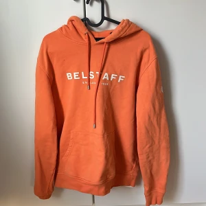 Orange hoodie från Belstaff - Säljer en snygg orange hoodie från Belstaff med vit logga på bröstet och ärmen. Tröjan har en klassisk känguruficka och justerbar huva med dragsko. Perfekt för en avslappnad stil. Pris går att diskutera 