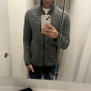 Grå fleecejacka från Patagonia - Säljer en grå fleecejacka från Patagonia med dragkedja framtill. Jackan har en normal passform och är perfekt för kyligare dagar. Den är långärmad och har en enkel design som passar till det mesta.