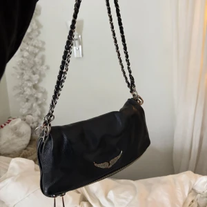 Svart axelväska från Zadig & Voltaire - Snygg svart axelväska i skinn från Zadig & Voltaire med kedjedetaljer på axelremmen. Väskan har en dragkedja och det medkommer 3 kedjor i olika längder (två av kedjorna har små defekter). Den är i bra skick men har fått brukslitage samt patina på silver detaljerna, finns även en liten defekt i högra hörnet därav priset! 