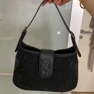 Svart axelväska med glittrig detalj - Snygg svart axelväska med glittrig framsida och stilren design. Väskan har en praktisk axelrem och en elegant stängning framtill. Perfekt för att ge en touch av glamour till din outfit.