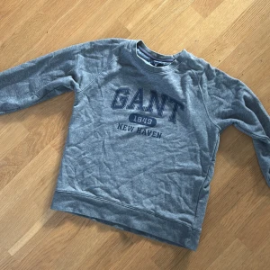 Grå sweatshirt från GANT - Säljer en grå sweatshirt från GANT med tryck 'GANT 1949 New Haven' på framsidan. Tröjan har långa ärmar och ribbade muddar vid ärmslut och nederkant. Perfekt för en avslappnad stil.