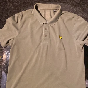 Olivgrön pikétröja med gul logotyp - Snygg olivgrön pikétröja från Lyle & Scott med klassisk krage och tre knappar. Tröjan har en gul logotyp på bröstet som ger en stilren touch. Perfekt för en avslappnad men ändå uppklädd look. Använd i 1 månad, men har ej kvitto.🌳