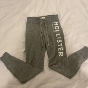 Grå mjukisbyxor från Hollister - Säljer ett par grå mjukisbyxor från Hollister med snörning i midjan och ribbade muddar. Perfekta för en avslappnad stil med tryck längs benet.