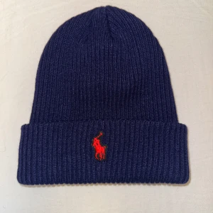 Navy blå Ralph Lauren mössa - Helt oanvänd Snygg mörkblå stickad mössa från Polo Ralph Lauren med röd broderad logga framtill. Perfekt för kyliga dagar och ger en stilren look. Tillverkad i tjockt och mjukt material för extra värme och komfort.                                Priset kan diskuteras vid snabb affär!