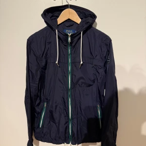 Polo Ralph Vindjacka - Snygg mörkblå vindjacka från Ralph Lauren med huva och dragkedja i grönt och vitt. Jackan har två fickor med dragkedja och en broderad logga på ärmen. Perfekt för blåsiga dagar! Pris kan diskuteras vid en snabb affär😃
