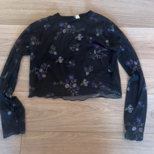 Svart blommig långärmad topp - Snygg svart långärmad topp med blommigt mönster i lila och grönt. Perfekt för en stilren look med en touch av natur. Toppen har en lätt croppad design med vågig nederkant.