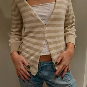 Beige och guldglittrig kofta - Snygg beige kofta med glittriga guldiga ränder. Den har en v-ringad design med trekvartslånga ärmar, perfekt för att lägga till lite glamour till din outfit.