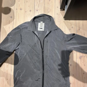 Grå jacka från Duno - Grå jacka eller overshirt från Duno i speciellt reflexmaterial. Väldigt tunn sommarjacka. Storlek M. Ett synligt märke finns på sista bilden. Nypris 2700