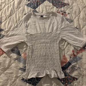 Vit blus med smockdetaljer - Säljer en söt vit blus med smockdetaljer och volangkant. Blusen har trekvartsärmar och är perfekt för en avslappnad stil. Passar bra till både jeans och kjol. Sömmen är sönder!