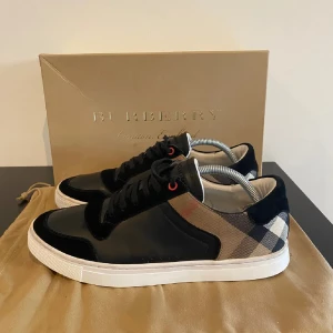 Svarta sneakers från Burberry - Stilrena svarta sneakers från Burberry med klassiskt rutigt mönster på sidorna. Skorna har snörning och en vit sula som ger en snygg kontrast. Perfekta för en trendig och elegant look.