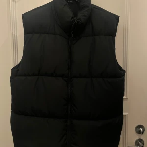 Svart dunväst från BikBok - Dunväst från BikBok i storlek L. Västen har en dragkedja framtill och justerbar dragsko i nederkant för perfekt passform. Fickor på sidan.